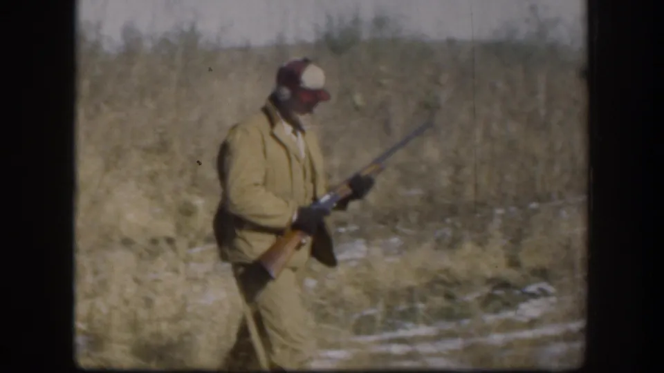 1958 Kansas home-movie frame of a hunter in tan clothing walking through grass with a rifle held downward.