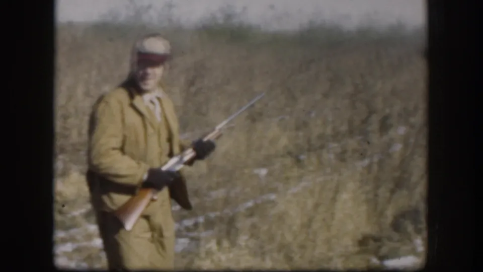 1958 Kansas frame of a man in a red cap looking toward the camera while holding a rifle in a grassy field.