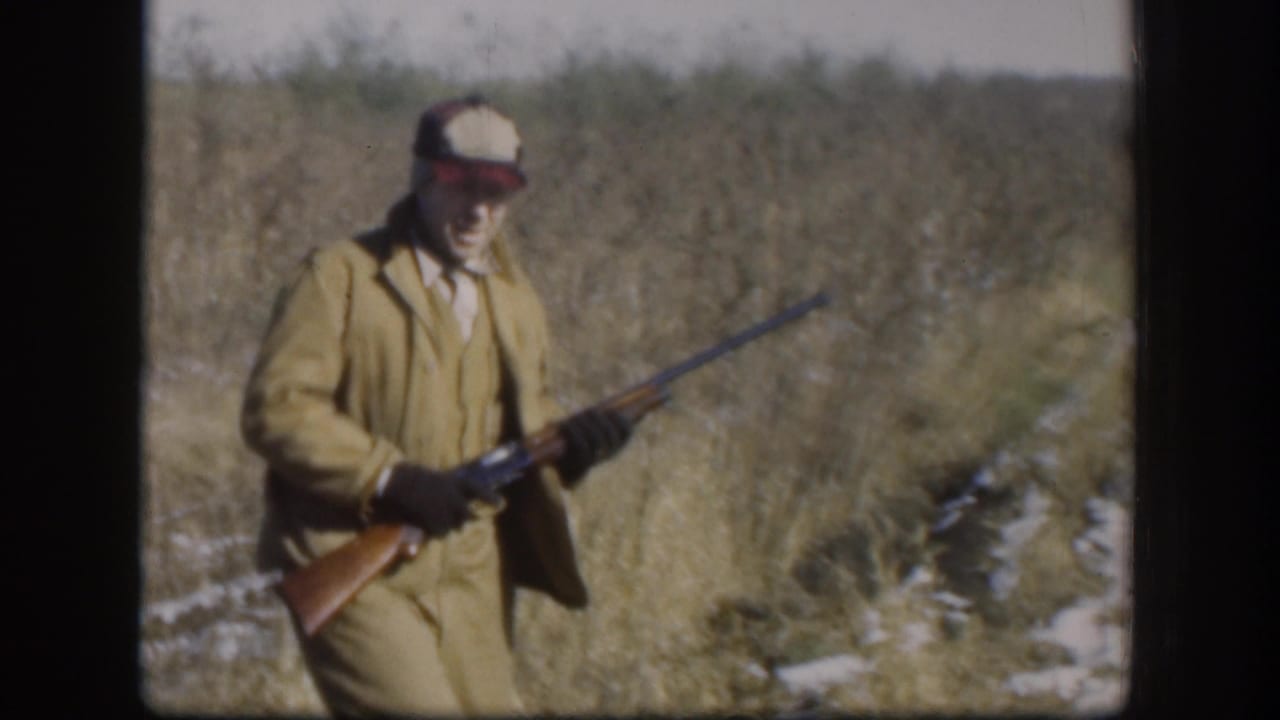 In Kansas 1958 home-movie footage, an adult hunter in tan field clothes carries a rifle through tall grass with snow patches.