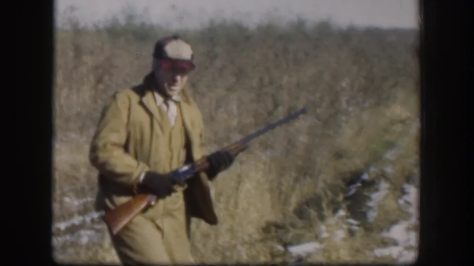 In Kansas 1958 home-movie footage, an adult hunter in tan field clothes carries a rifle through tall grass with snow patches.