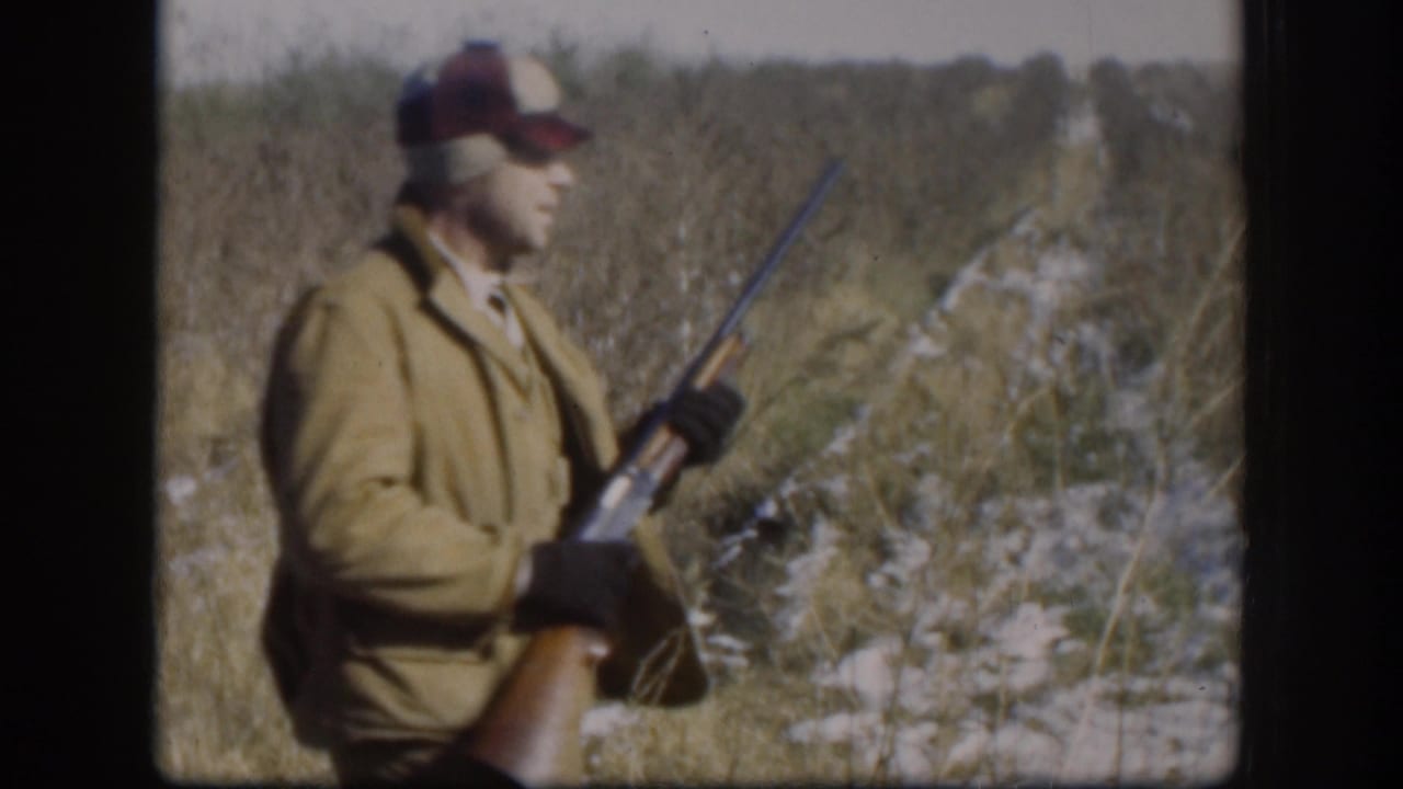 Hunter stands in profile in tall grass, holding a rifle across his body.