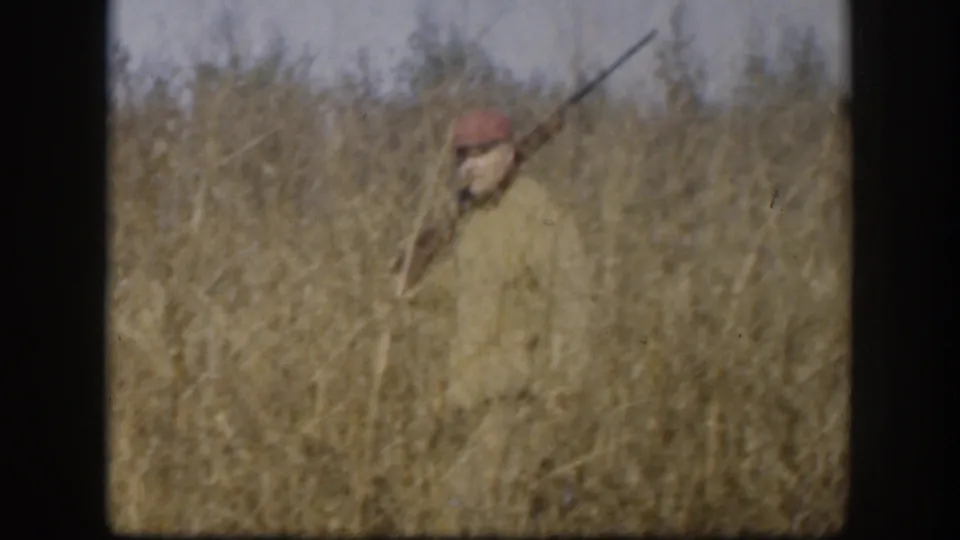 The hunter appears partly obscured by tall Kansas grass as he raises or aims a rifle in the 1958 home-movie frame.