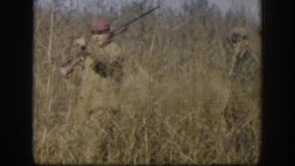 An adult hunter leans through tall grass with a rifle extended, seen in soft 8mm Kansas home-movie footage from 1958.