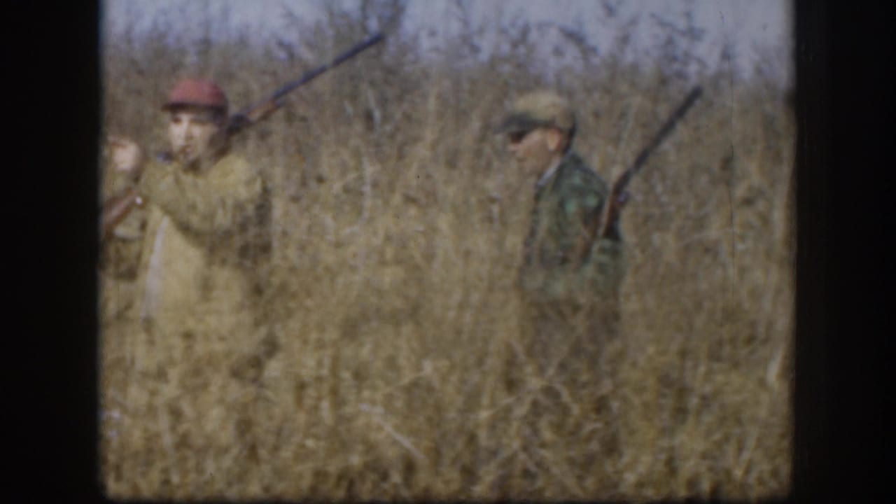 Two hunters in caps stand in dry Kansas brush, one in a red cap and another in a plaid shirt with a rifle.