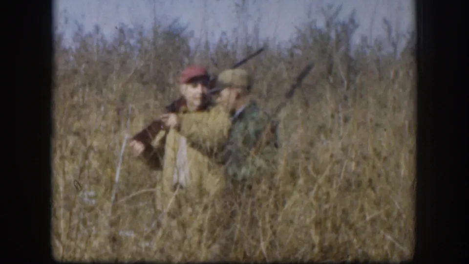 A red-capped hunter and a man in a green plaid shirt stand close together in a 1958 Kansas field, both carrying rifles.