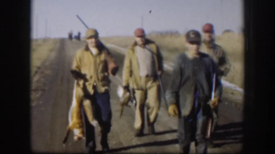 A group of men walk toward the camera on a rural Kansas road in 1958, with rifles and a carried fox visible.