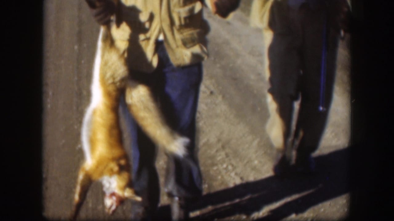 Hunter holding a dead fox by its legs beside another man on a dirt path.