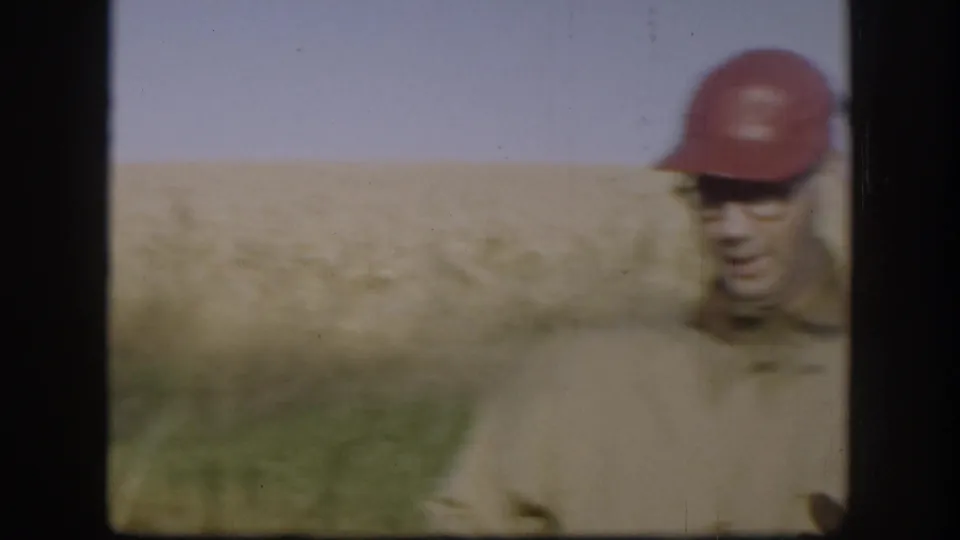 Soft 8mm frame of an adult man in a red cap near the right edge, with rural grassland behind him.