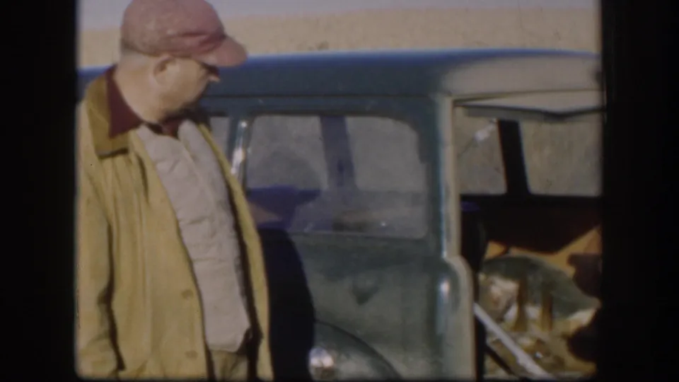 Adult man in a red cap and light jacket stands beside an open green car in rural Kansas footage from 1958.
