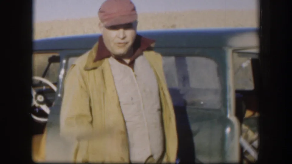 Adult man in a red cap faces the camera near a green vintage car in 1958 Kansas home-movie footage.