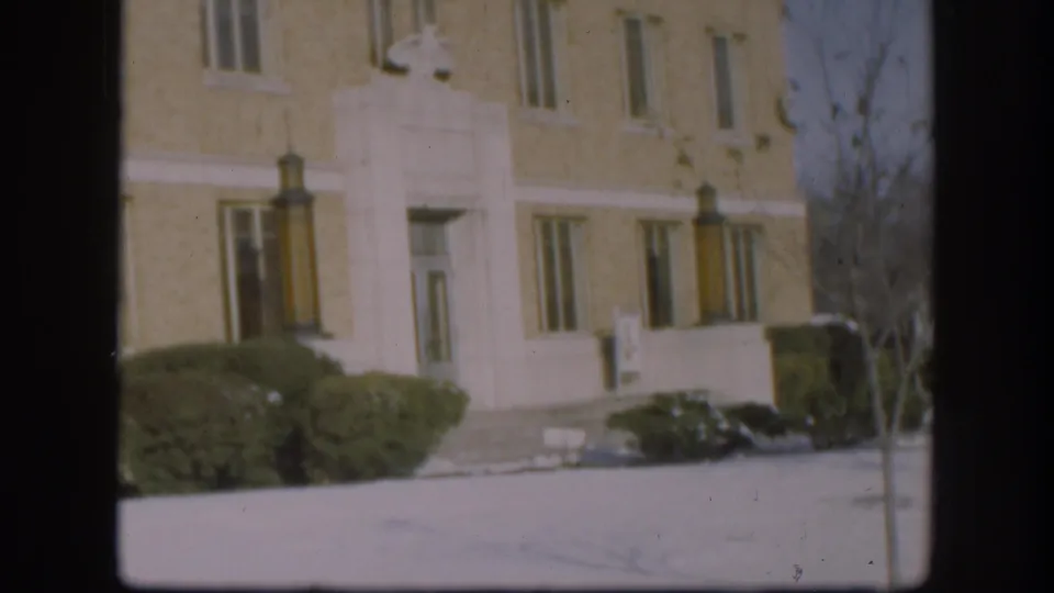 1958 Kansas home-movie frame showing the Clark County Courthouse entrance, light brick facade, white trim, shrubs, and snow on the grounds.