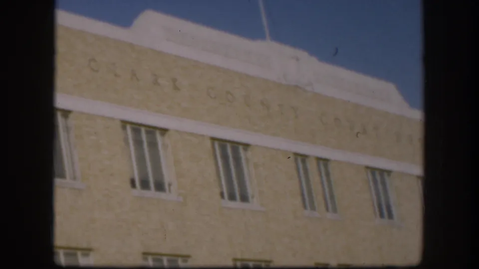 Handheld 1958 Kansas view of the Clark County Courthouse upper facade, showing light brick, white roofline trim, windows, and lettering.