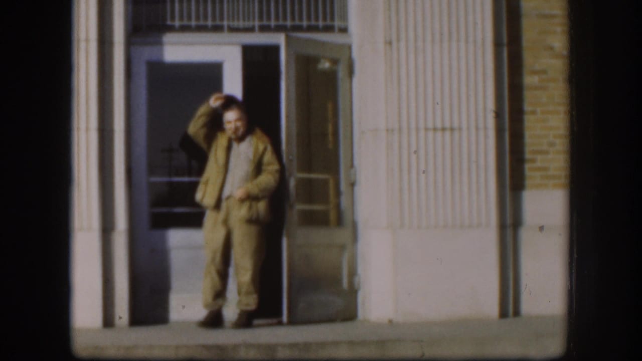 Man in tan work clothes stands in a doorway, holding a cigar near his head.