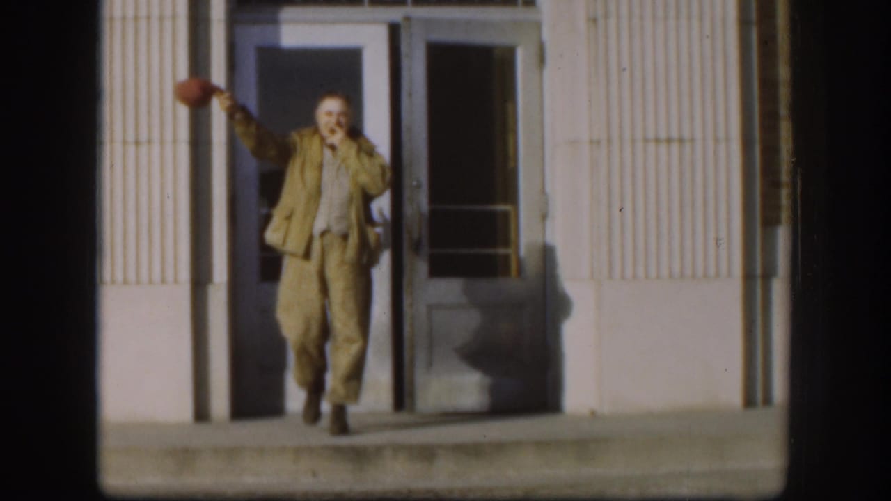 In the Kansas 1958 home-movie frame, the man smiles broadly and waves a red cap from the building entrance.