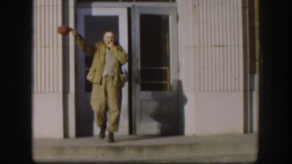 In the Kansas 1958 home-movie frame, the man smiles broadly and waves a red cap from the building entrance.