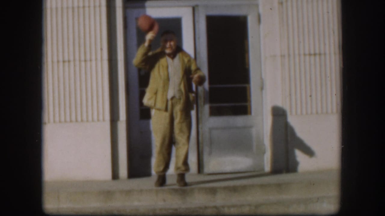 The man faces the camera in tan workwear, holding his red cap near his head and a cigar in the other hand.
