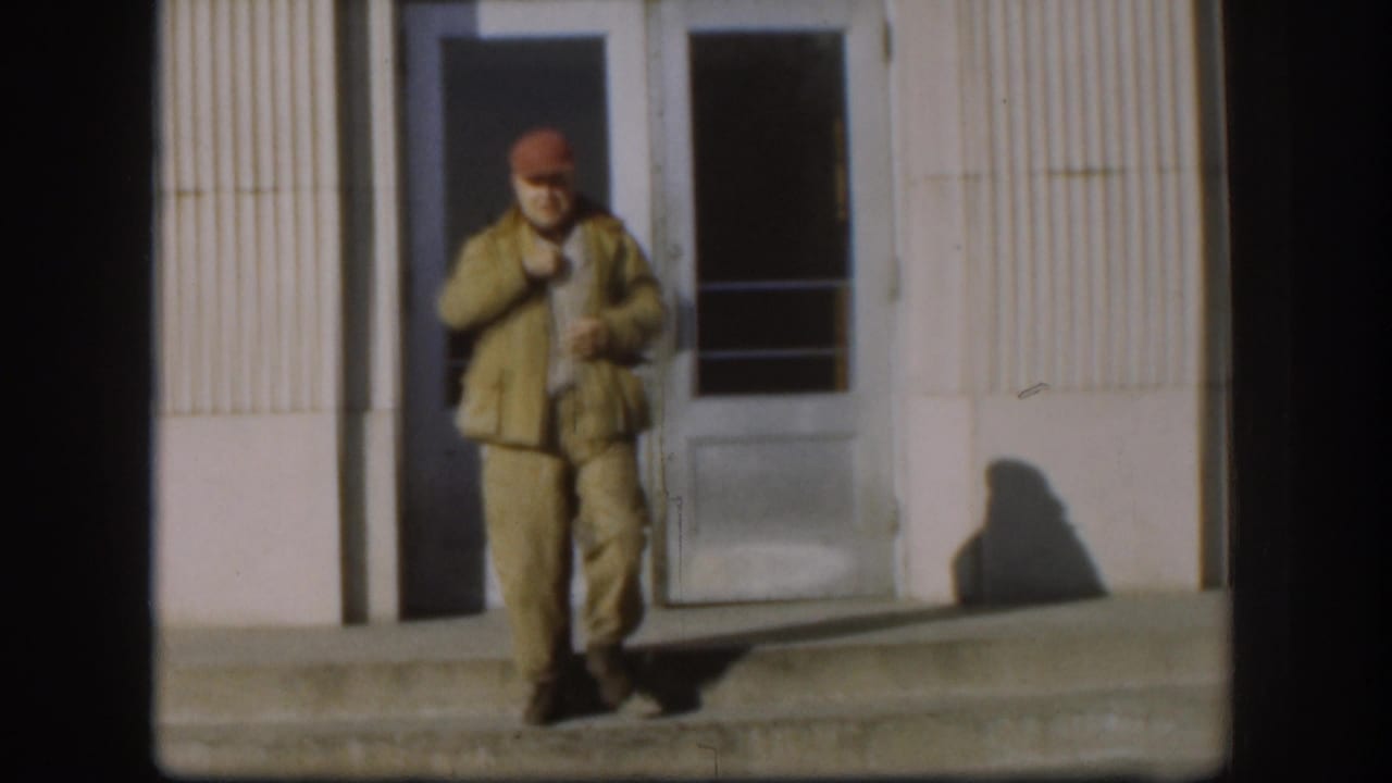 The man stands near the doorway in bright sun, holding a cigar while his shadow falls across the building wall.