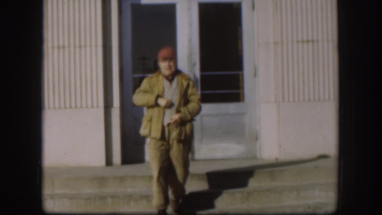 Man in a red cap and tan jacket stands near steps outside double doors.