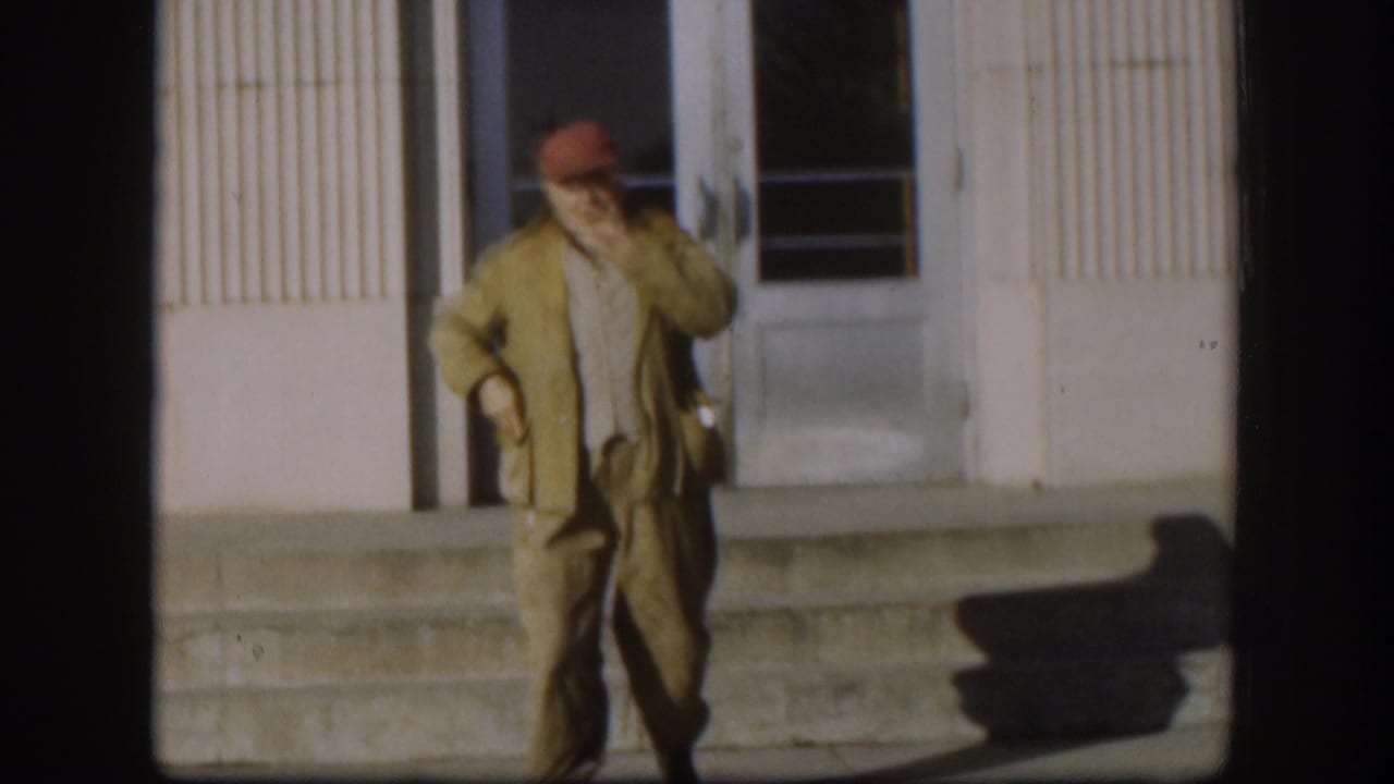 The adult man in a red cap and tan jacket walks down steps outside the Kansas building, raising a cigar near his face.
