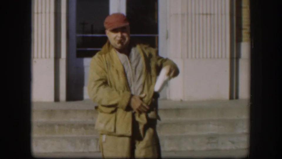 In the final Kansas 1958 still, the man stands closer to camera, handling a small item near his jacket with the building behind him.