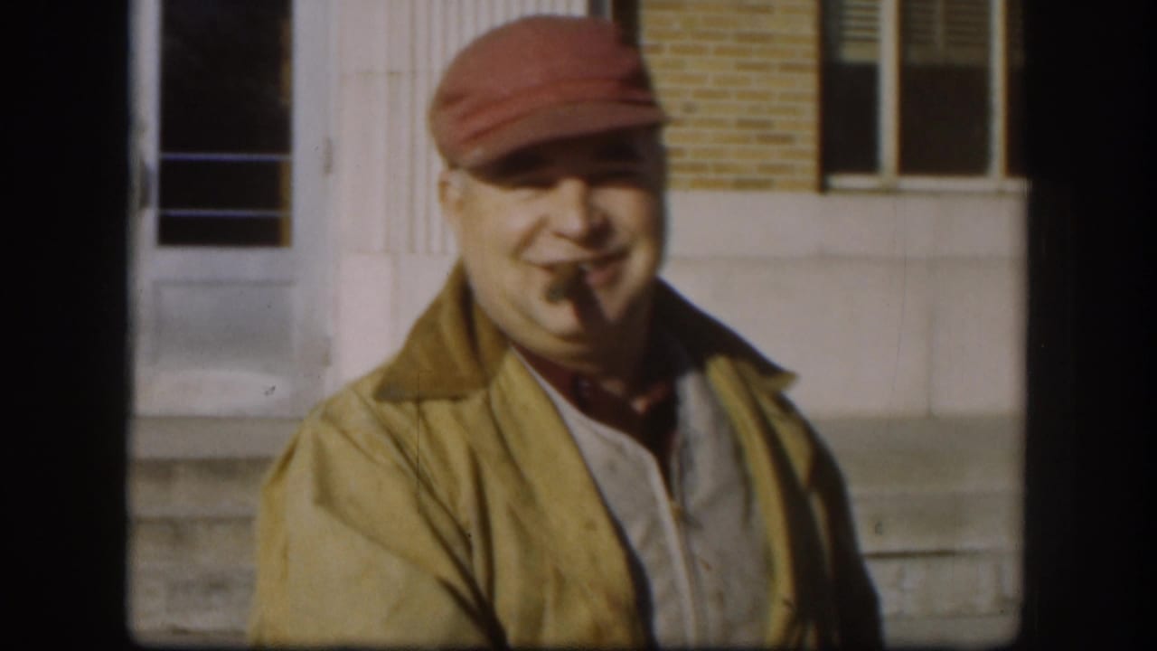 In Kansas 1958 footage, an adult man in a red cap and yellow jacket smiles outdoors near a brick building while holding a cigar in his mouth.