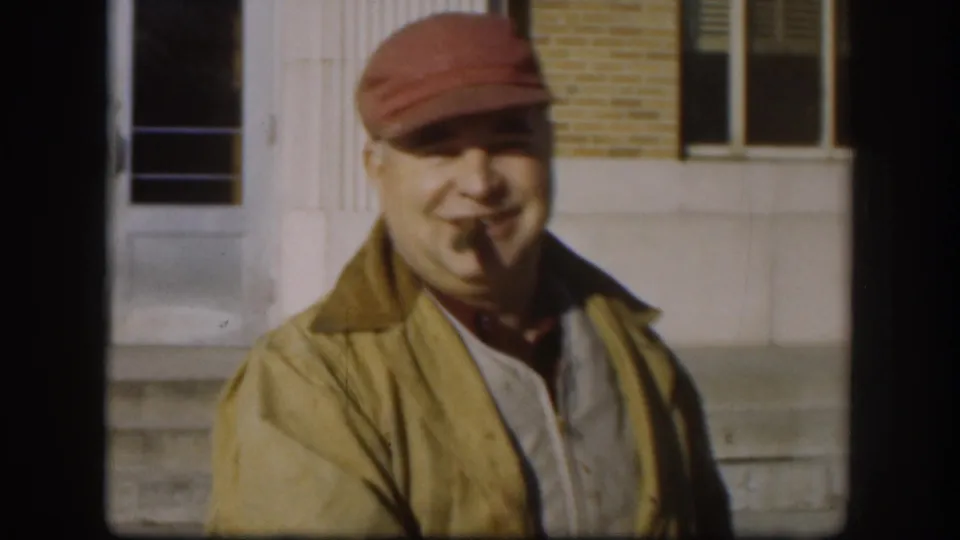 In Kansas 1958 footage, an adult man in a red cap and yellow jacket smiles outdoors near a brick building while holding a cigar in his mouth.