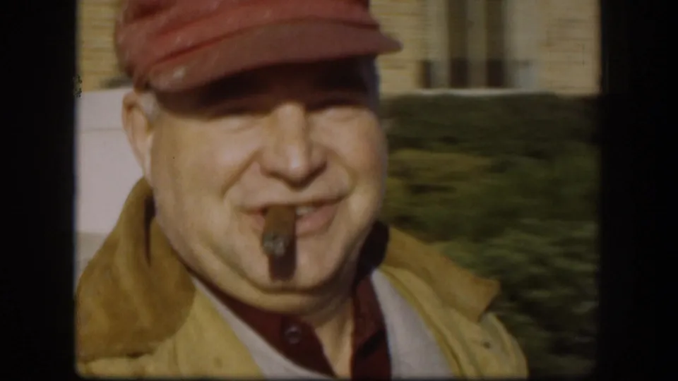 Close 1958 Kansas home-movie view of a smiling adult man in a red cap and yellow jacket with a cigar in his mouth.