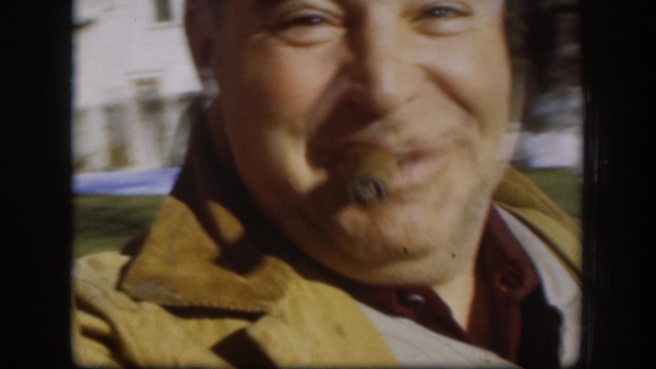 A very close Kansas 1958 frame shows the adult man smiling into the camera, with a cigar, red cap brim and yellow jacket visible.