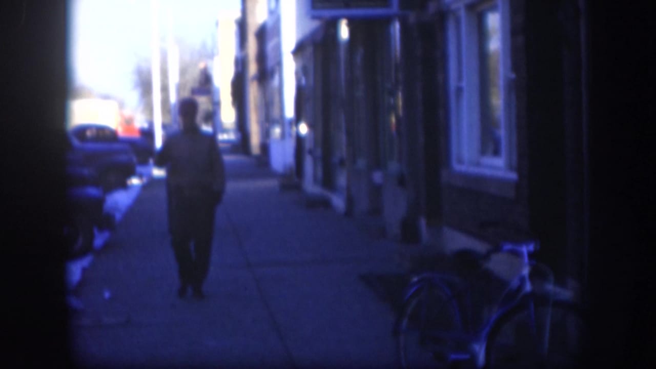 Blue-tinted 1958 Kansas street frame showing a person walking away on a sidewalk beside parked cars, building fronts and a bicycle.