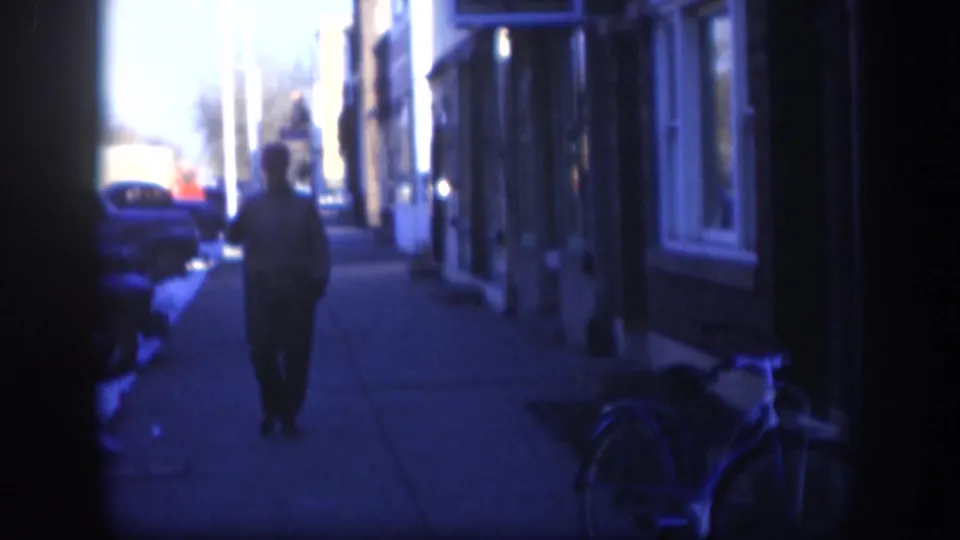 Blue-tinted 1958 Kansas street frame showing a person walking away on a sidewalk beside parked cars, building fronts and a bicycle.