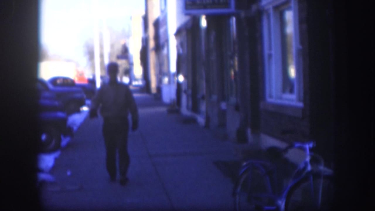 1958 Kansas home-movie frame showing a man in a cap on a snowy sidewalk, with parked cars, storefront windows, and a bicycle at the right edge.