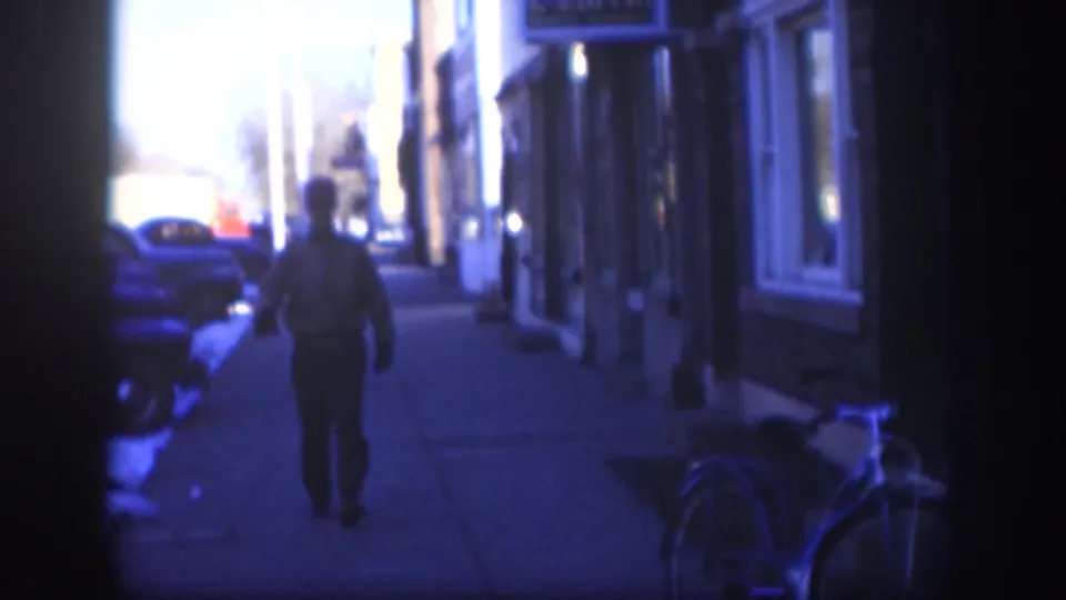 1958 Kansas home-movie frame showing a man in a cap on a snowy sidewalk, with parked cars, storefront windows, and a bicycle at the right edge.