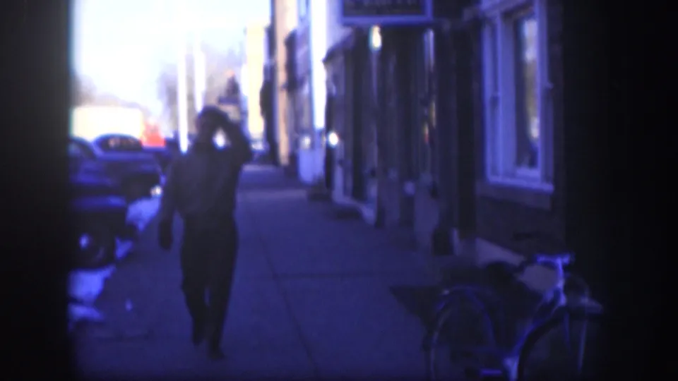 1958 Kansas street frame with a man raising his hand to his cap as he walks on a blue-cast snowy sidewalk past cars and shopfronts.