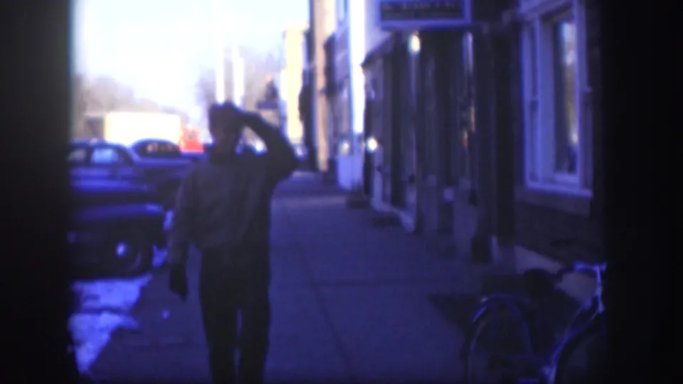 The man moves closer in the 1958 Kansas sidewalk scene, hand still near his cap, with vintage cars, storefronts, and a bicycle visible around him.