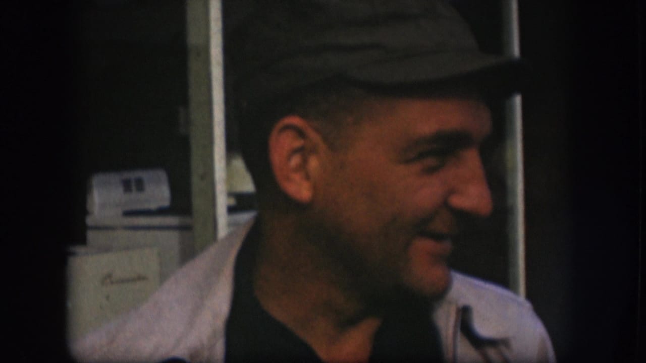 Man in a dark cap and light jacket smiles beside a window or door frame.