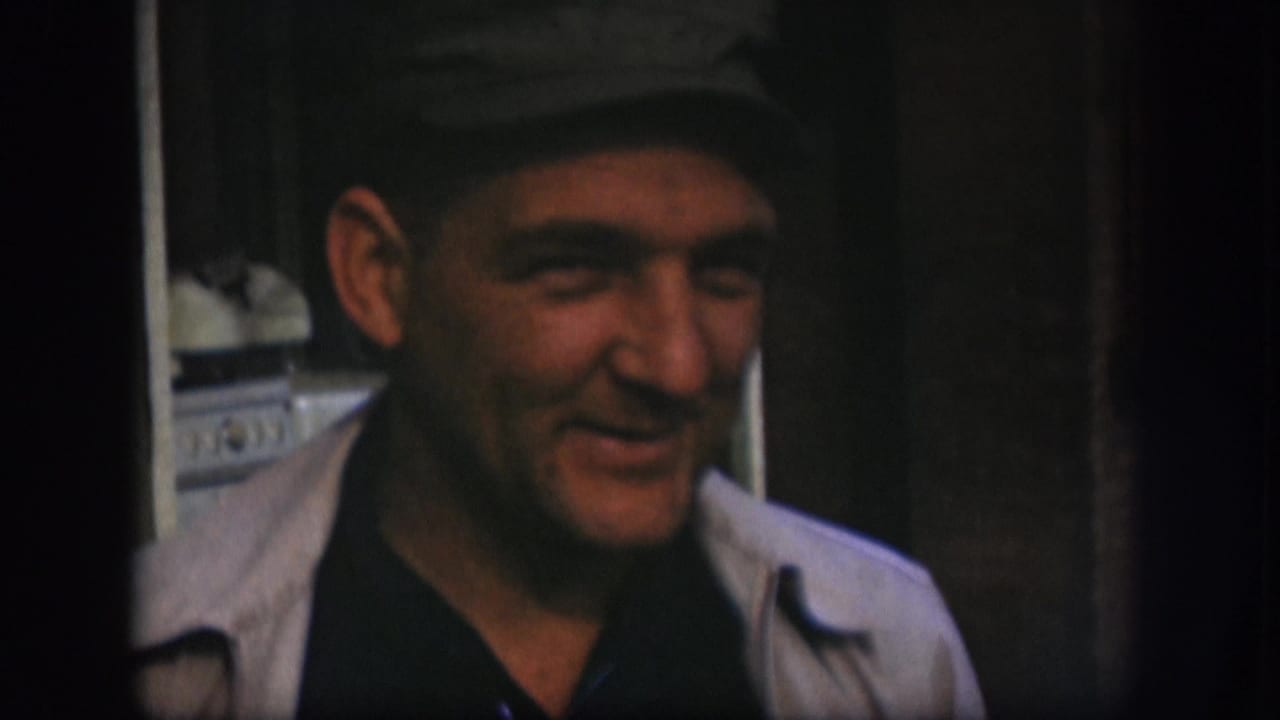 The cap-wearing man laughs in a 1958 Kansas interior, with wood trim and a pale appliance partly visible behind him.