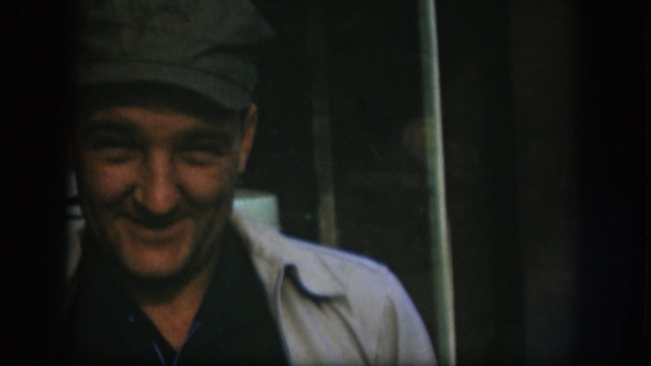 The man smiles downward in a 1958 Kansas interior, framed by dark edges and a vertical window or door element.
