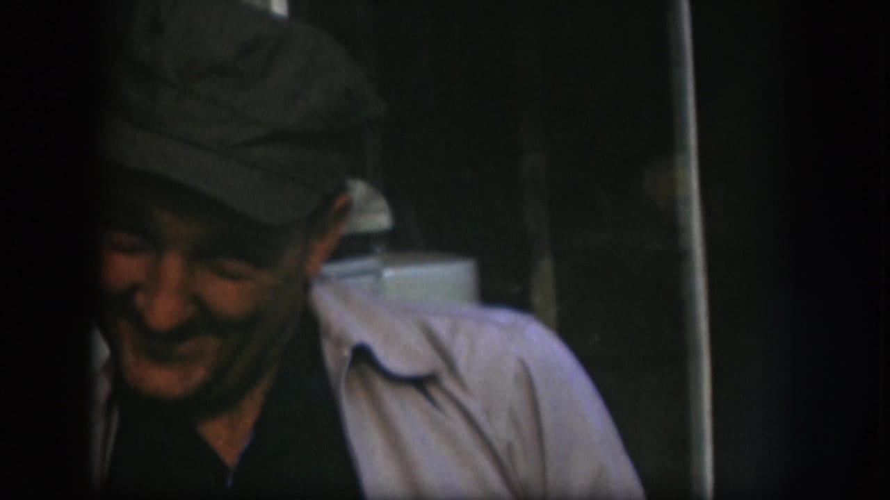 Older man in a dark cap and light jacket smiles near a doorway in Kansas 1958 home-movie footage.