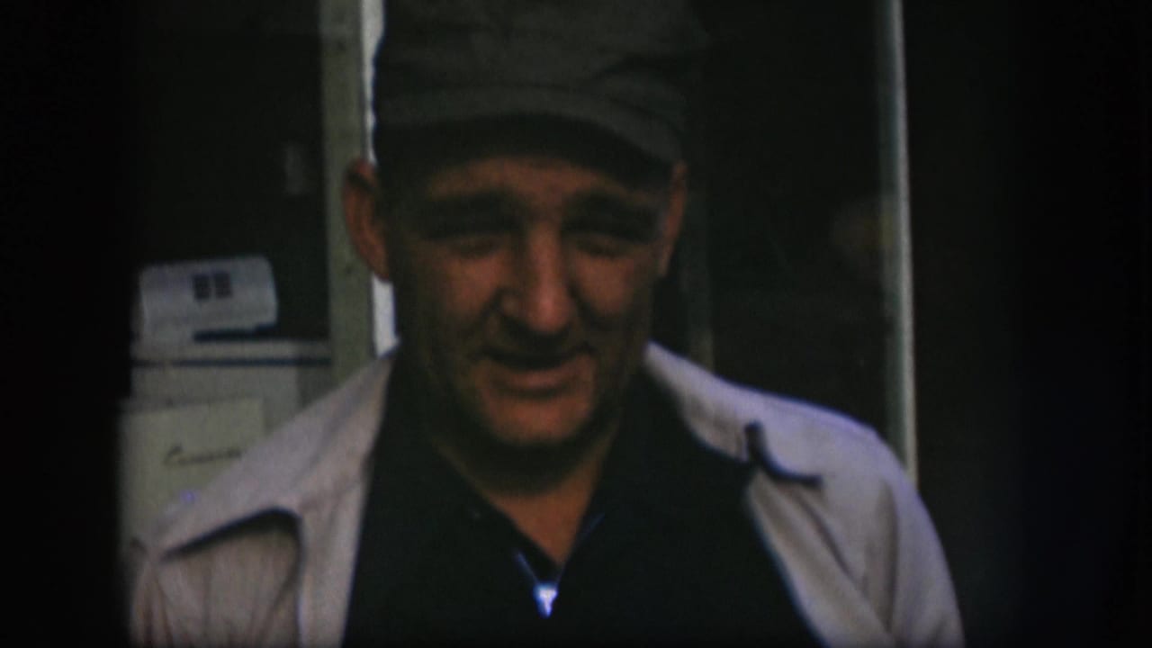 Older man in a cap and light jacket faces the camera, with a white appliance visible behind him in the interior.