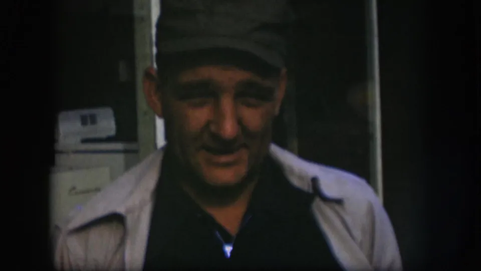 Older man in a cap and light jacket faces the camera, with a white appliance visible behind him in the interior.
