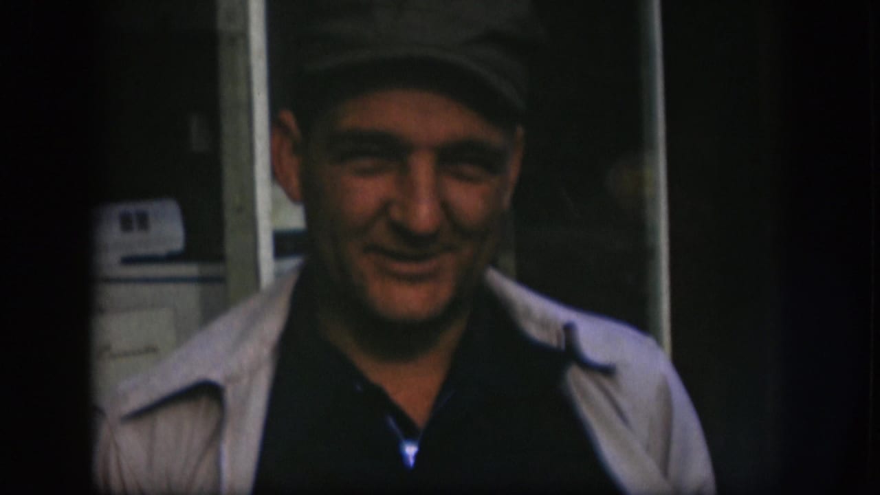 Kansas 1958 home-movie frame of an older man close to the camera, wearing a cap and light jacket in a dim room.