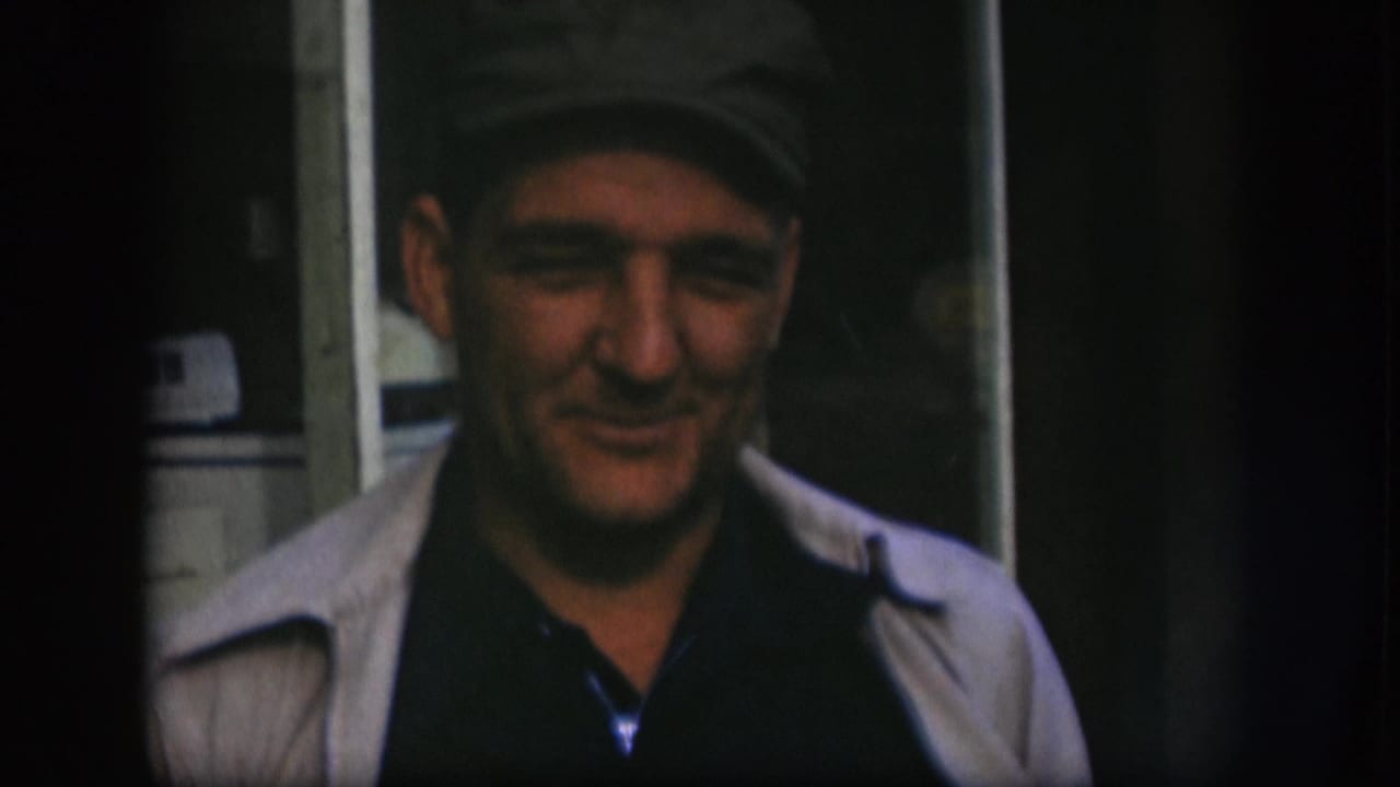 Older man in a dark cap appears to speak toward the camera in a shadowed Kansas home-movie interior from 1958.