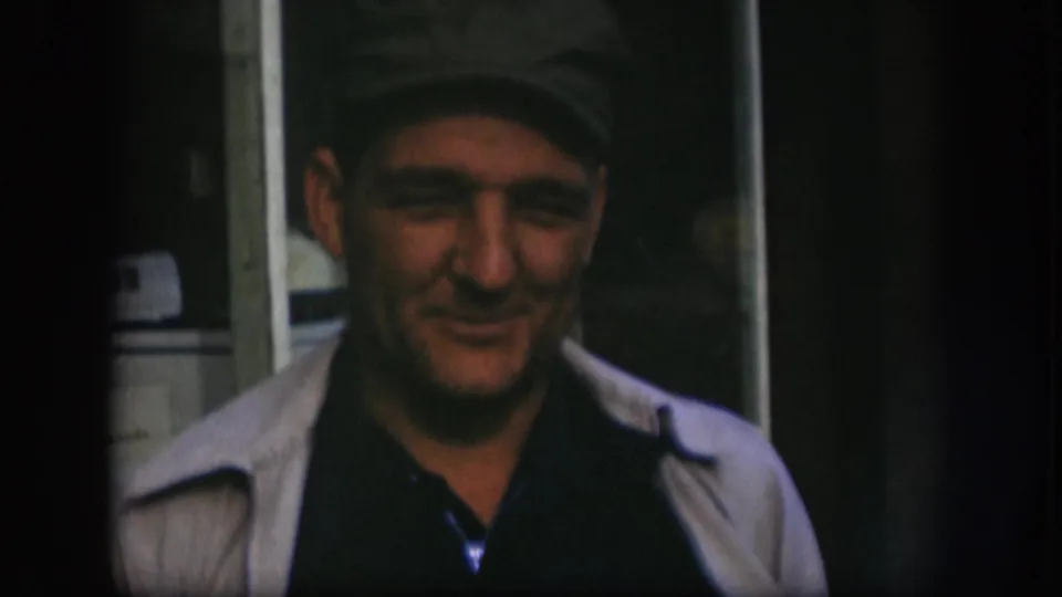 Older man in a dark cap appears to speak toward the camera in a shadowed Kansas home-movie interior from 1958.