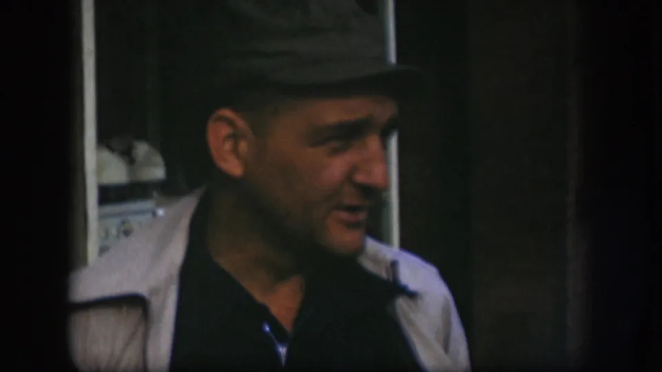 Kansas 1958 home-movie frame shows the man in side view, wearing a dark cap, dark shirt, and light jacket.