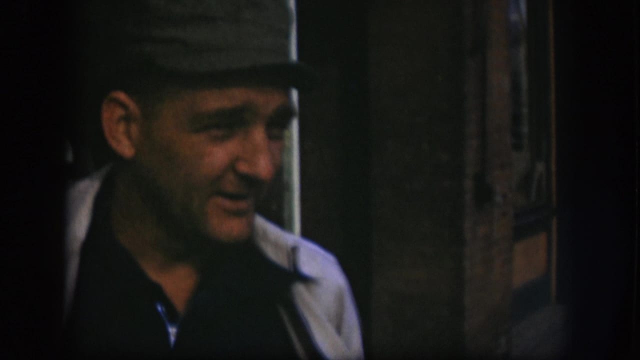 Adult man in 1958 Kansas 8mm footage wears a dark cap, dark shirt, and light coat in a dim interior setting.