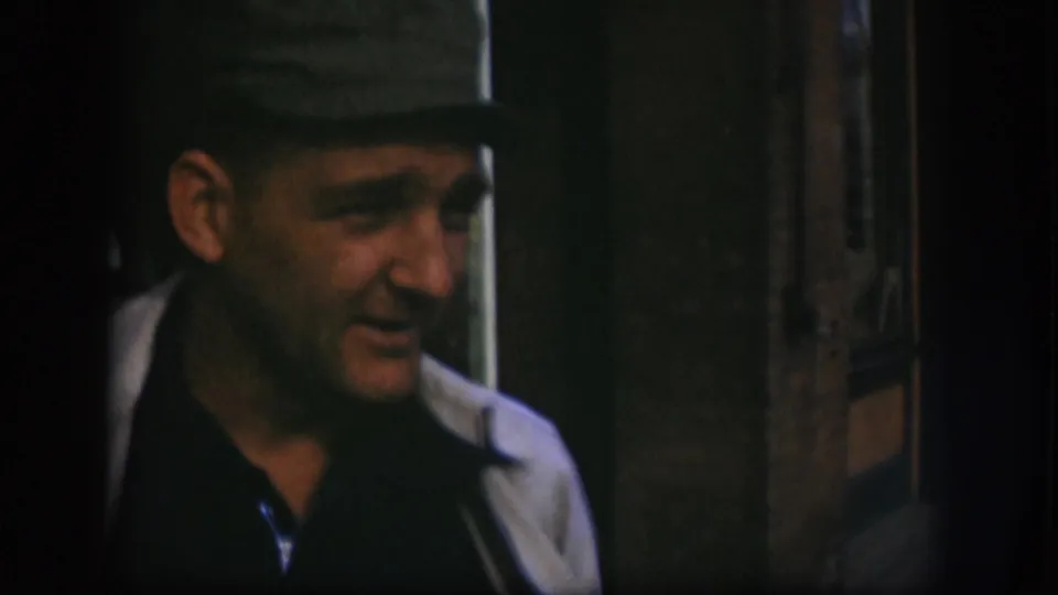 Adult man in 1958 Kansas 8mm footage wears a dark cap, dark shirt, and light coat in a dim interior setting.