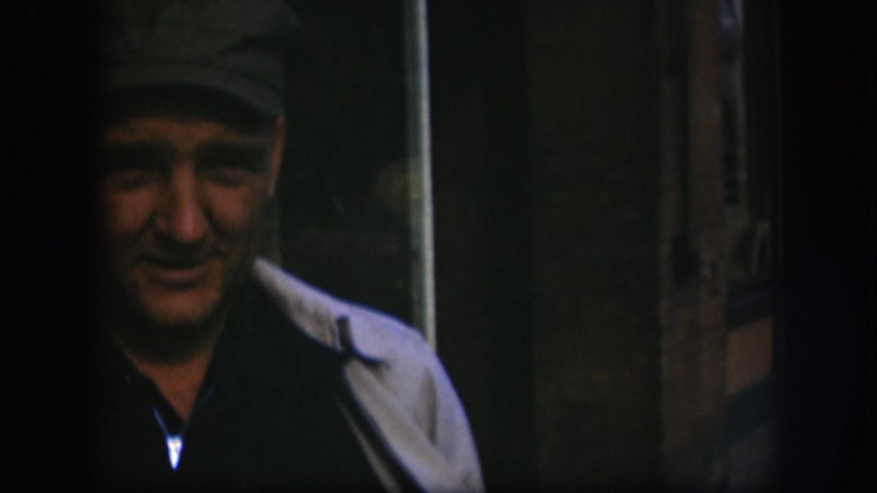 The man faces forward in soft 8mm color, with his cap and light coat visible against a dark Kansas interior.
