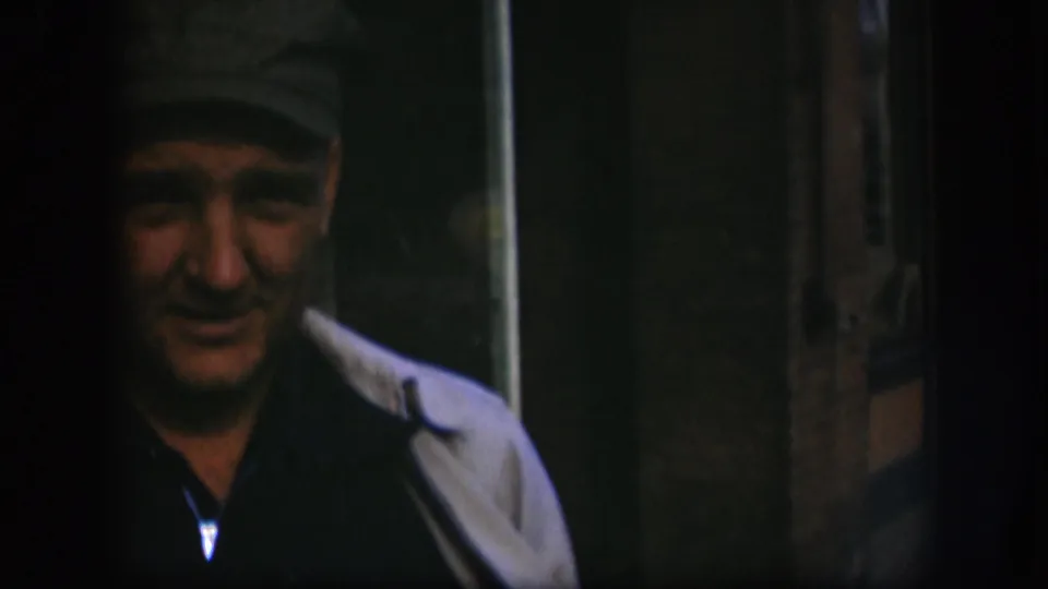 The man faces forward in soft 8mm color, with his cap and light coat visible against a dark Kansas interior.