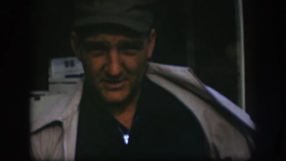 Adult man in a cap and light-colored coat faces the camera in dim 1958 Kansas 8mm footage.