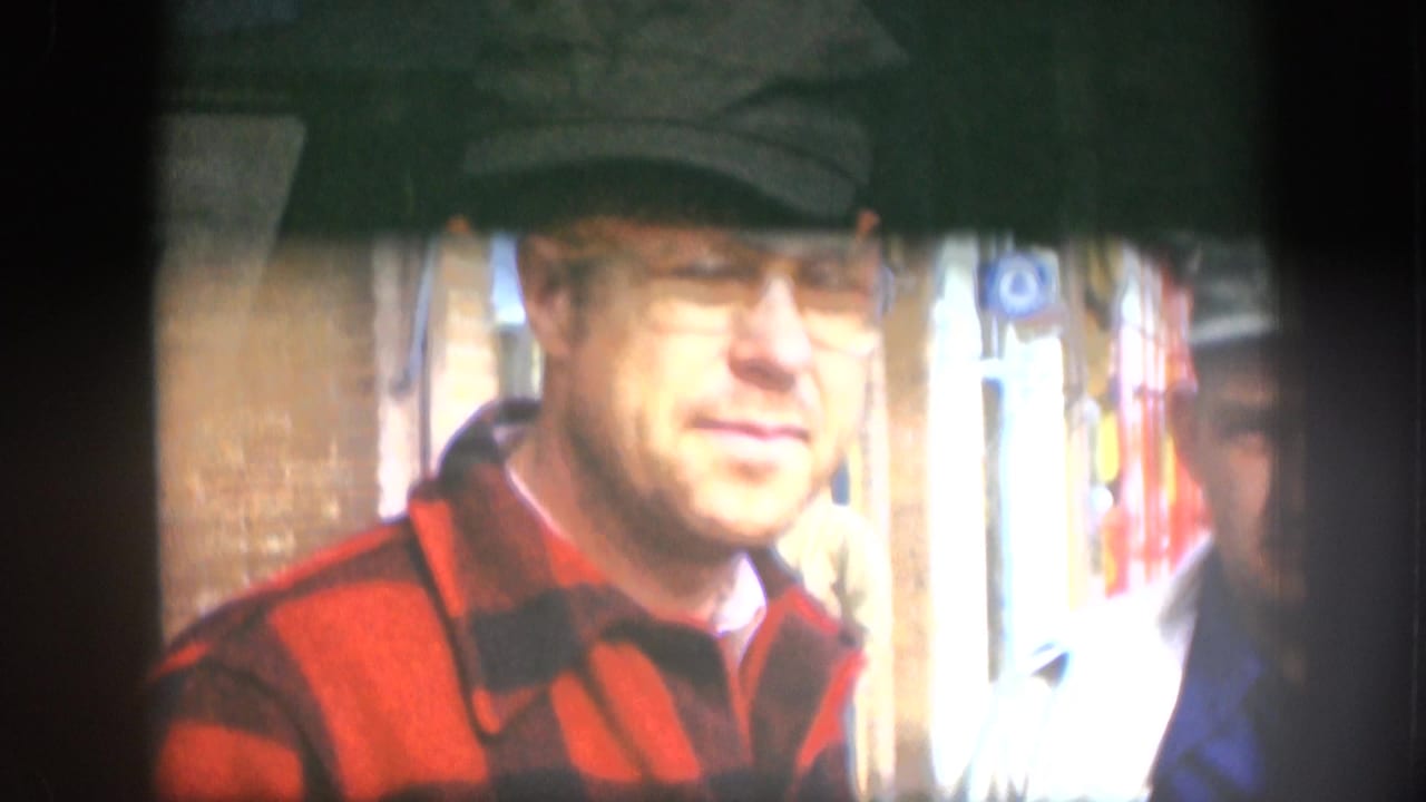 Adult man wearing glasses, a cap, and a red plaid jacket stands on a Kansas sidewalk in 1958 home-movie footage.